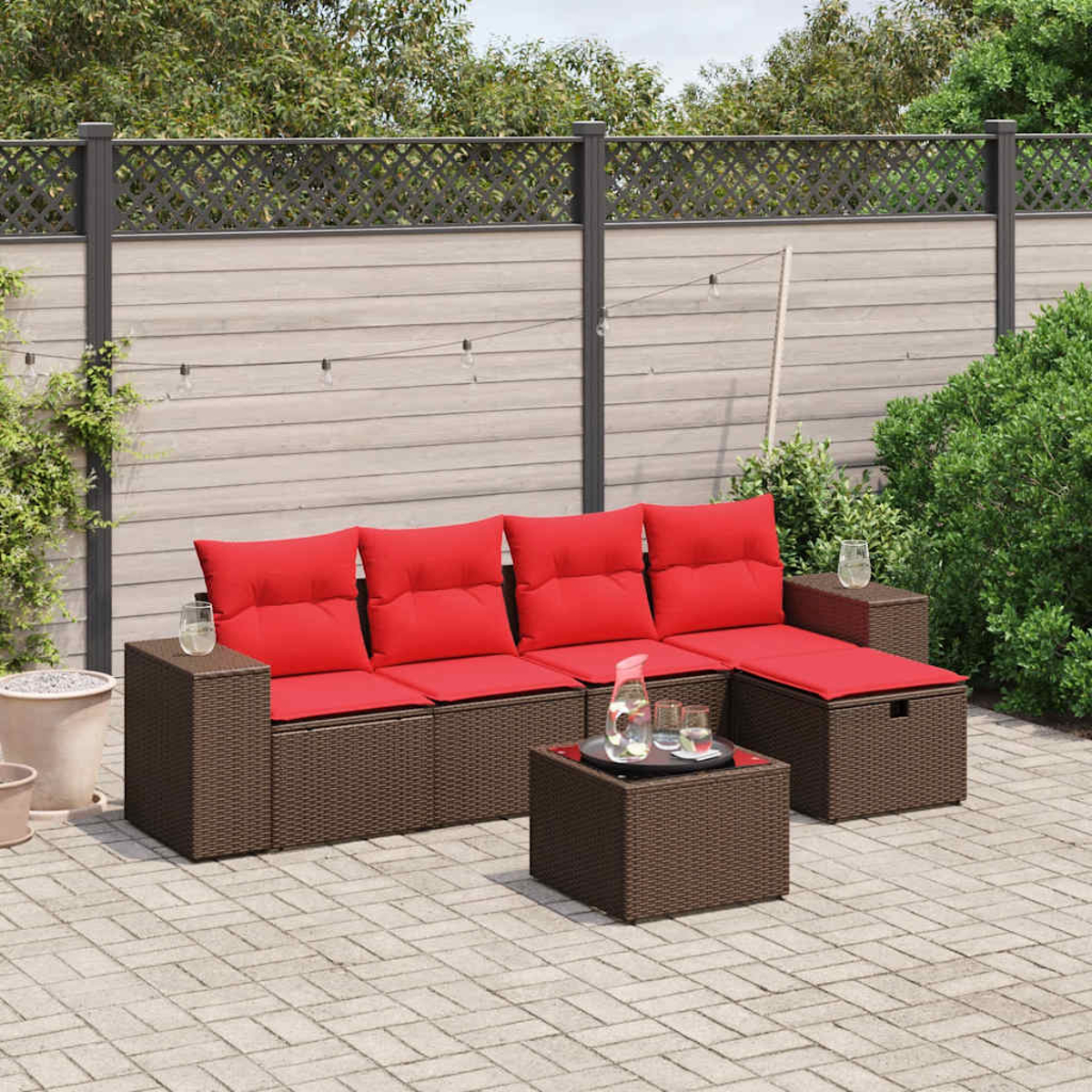 Garten-Sitzgruppe mit roten Kissen auf einem braunen Korbgewebe, kleinem Glastisch und einer Kulisse aus Holzzaun und Grün.