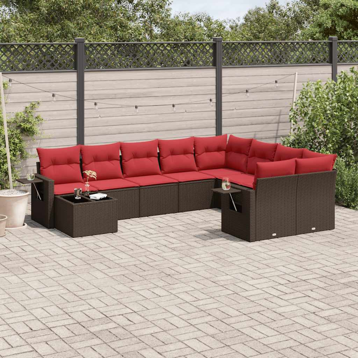Ein Ecksofa im Freien mit roten Kissen und braunem Korbgewebe. Es steht auf einer gepflasterten Terrasse mit Zaun und Pflanzen im Hintergrund.
