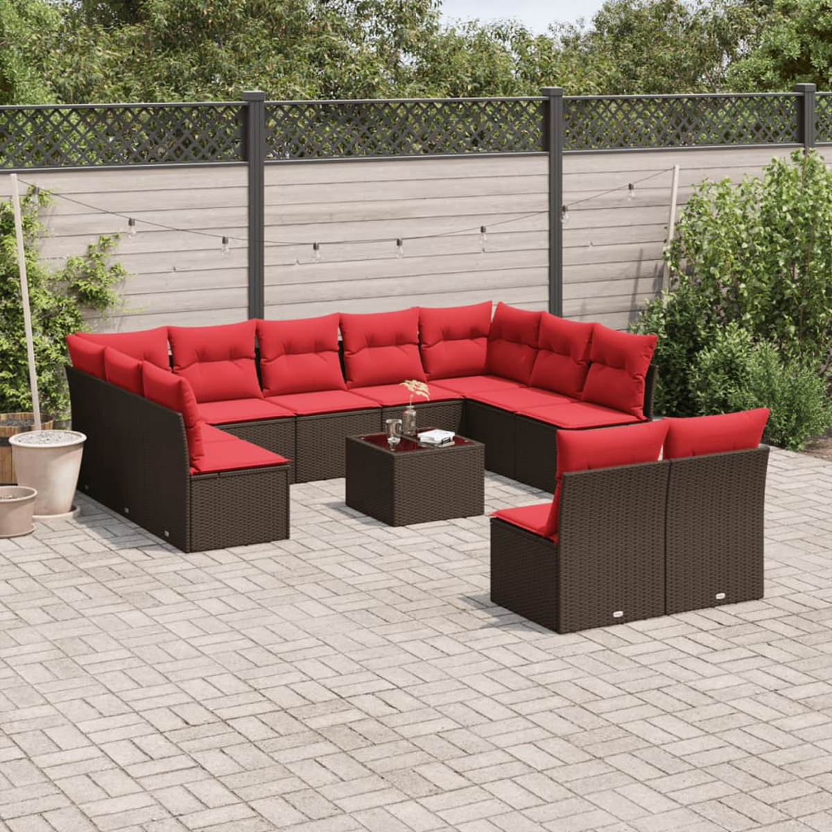 Gartencouch mit roten Kissen. Ein kleiner quadratischer Tisch und zusätzliche Sitzgelegenheiten sind ebenfalls zu sehen. Der Hintergrund zeigt einen Zaun und Grün.