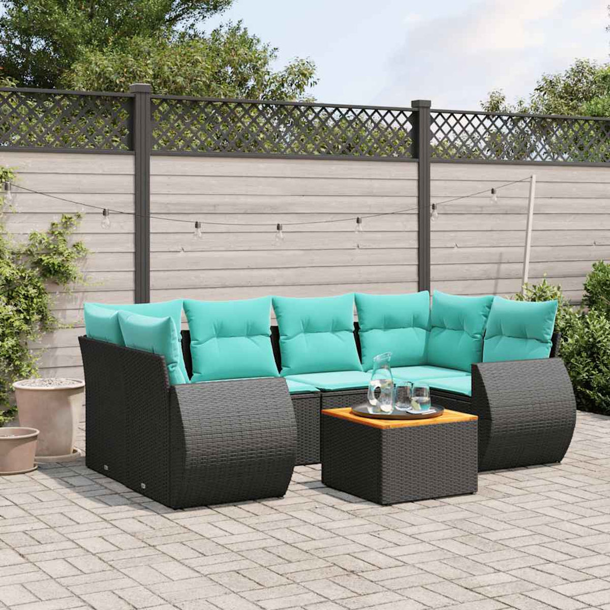 Garten-Lounge-Set mit schwarzem Webmöbel, türkisfarbenen Kissen und einem kleinen Holztisch. Ein Zaun und Pflanzen im Hintergrund.