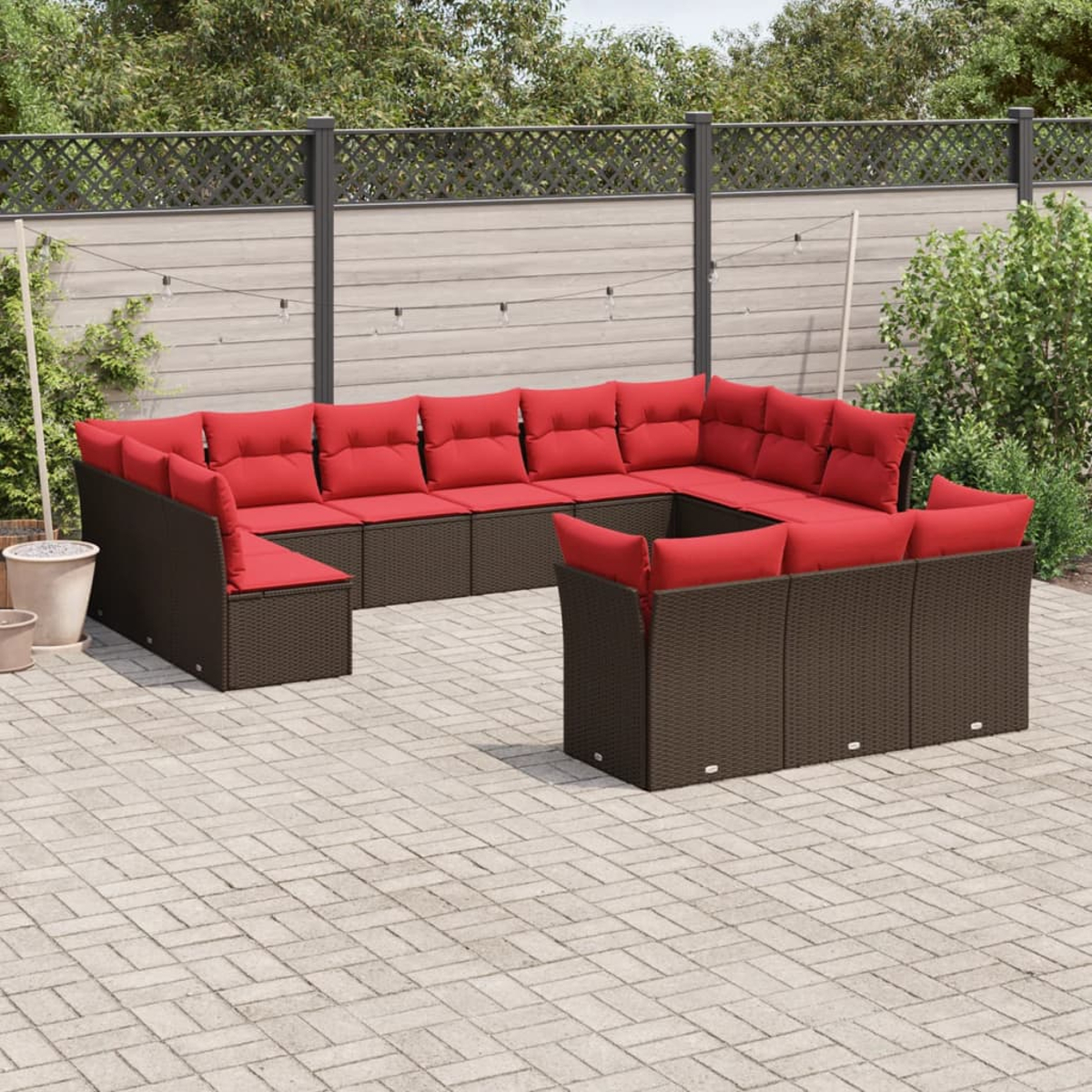Ein braunes Ecksofa aus Korbgeflecht mit roten Kissen steht auf einer Backstein-Terrasse. Im Hintergrund befinden sich ein Zaun und Grün.