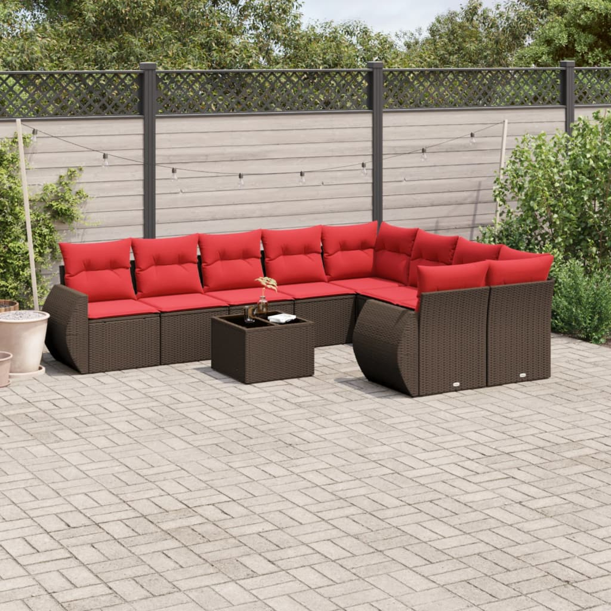 Gartenmöbel-Set mit roten Kissen. Braunes Korbmöbel-Ecksofa mit passendem Couchtisch auf einer Ziegelsteinterrasse mit Zaun.
