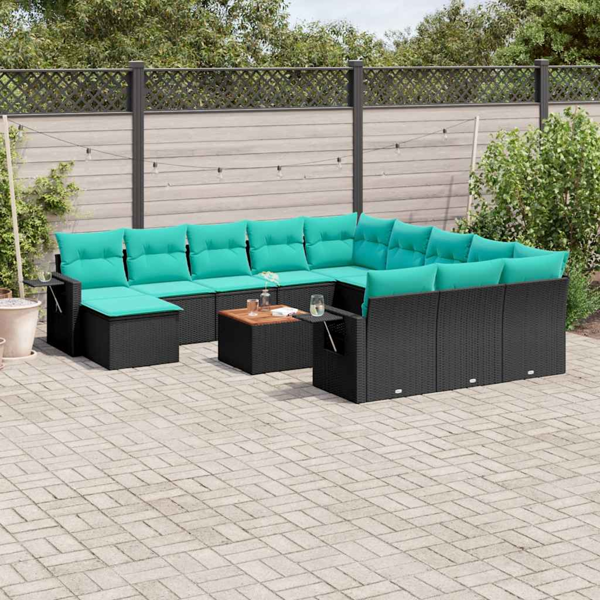 Ein schwarzes Outdoor-Ecksofa aus Korbgeflecht mit türkisfarbenen Kissen steht auf einer Terrasse. Ein kleiner Tisch hält Getränke bereit. Im Hintergrund befinden sich ein Zaun und Grün.