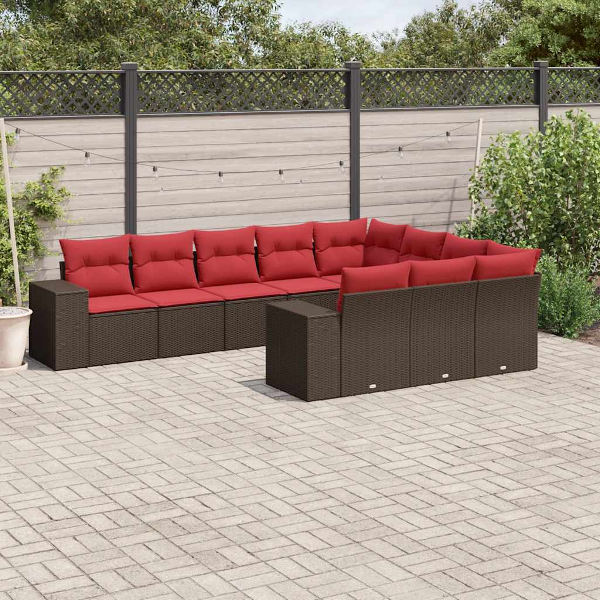 Outdoor-Ecksofa mit roten Kissen. Wicker-Style brauner Rahmen auf einer Stein-Terrasse. Zaun und Grün im Hintergrund.
