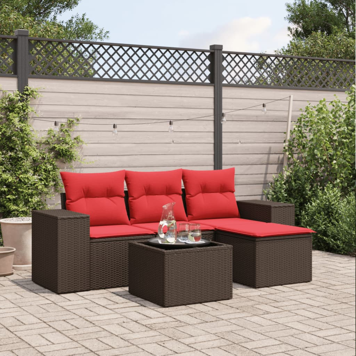 Garten-Terrassengarnitur mit roten Kissen, dunkelbraunem Korbgeflecht und einem Glastisch. Im Hintergrund ein Zaun und Pflanzen.