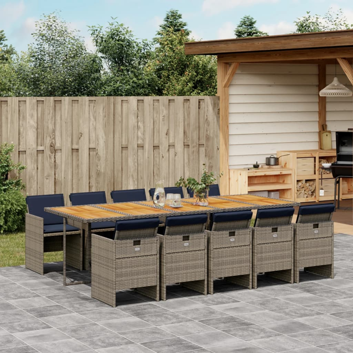 Essgruppe im Freien. Holztisch und Stühle mit blauen Kissen. Bäume und Holzzaun im Hintergrund, mit Terrasse.