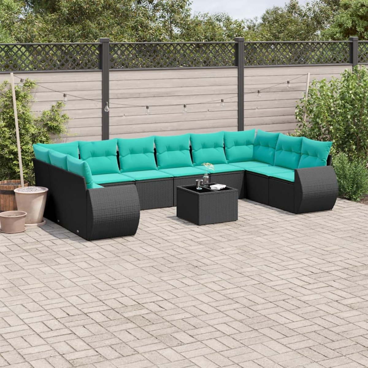 Outdoor-Ecksofa mit türkisfarbenen Kissen. Schwarzes Korbgeflecht mit passendem Couchtisch. Auf einer Backstein-Terrasse, mit Grün im Hintergrund.