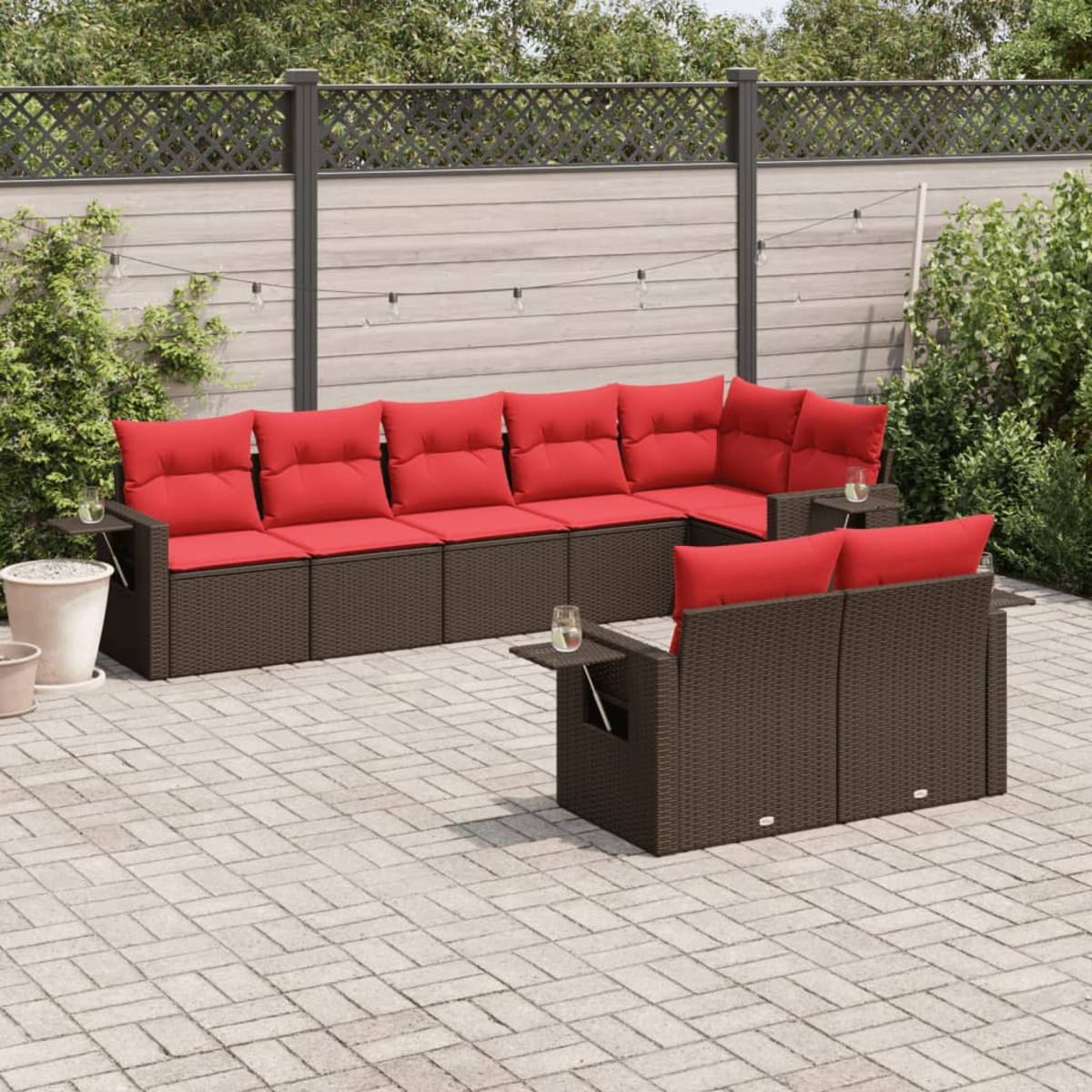 Terrassenset im Freien. Braunes Korbmöbel mit roten Kissen. Ein Tisch und ein Hintergrund aus Zaun und Grün.
