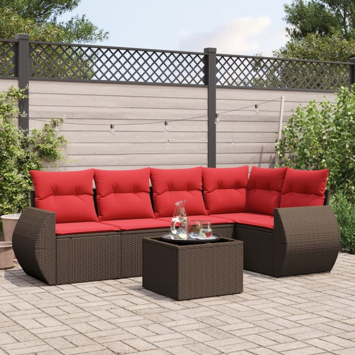 Garten-Terrassenset mit braunem Korbssofa mit roten Kissen und passendem Tisch. Im Hintergrund ein Zaun und Grün.
