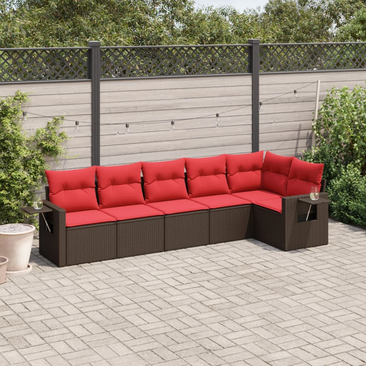 Ein Ecksofa mit roten Kissen im Freien steht auf einer Ziegelterrasse. Ein Holzzaun und Grünzeug sind im Hintergrund.