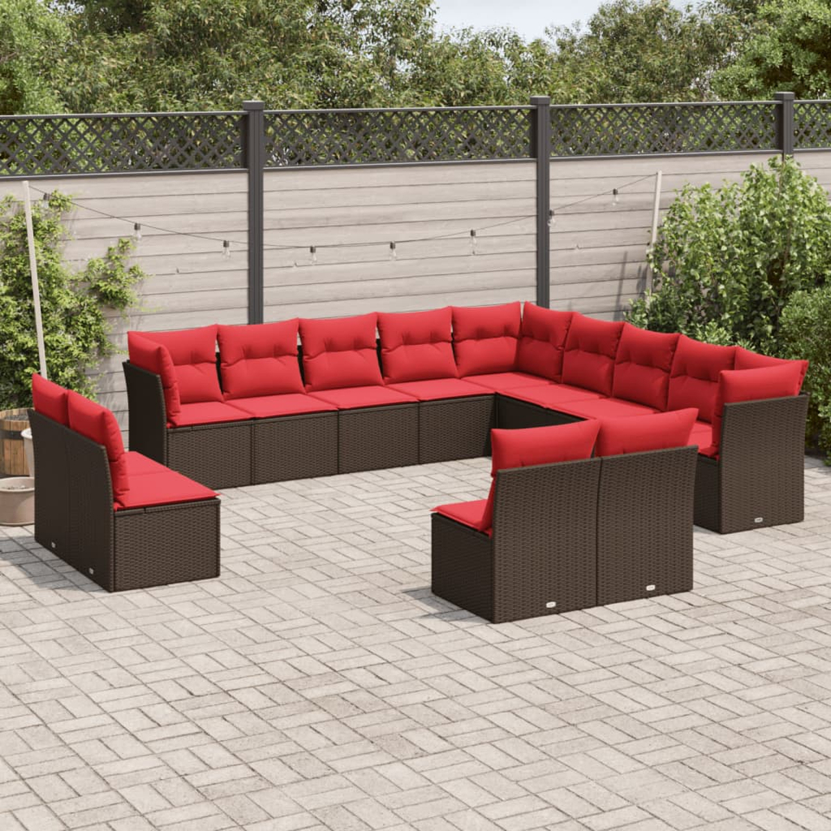 Ein Outdoor-Ecksofa mit roten Kissen steht auf einer Terrasse. Das Sofa ist aus braunem Korbgeflecht, der Hintergrund zeigt Bäume und einen Zaun.
