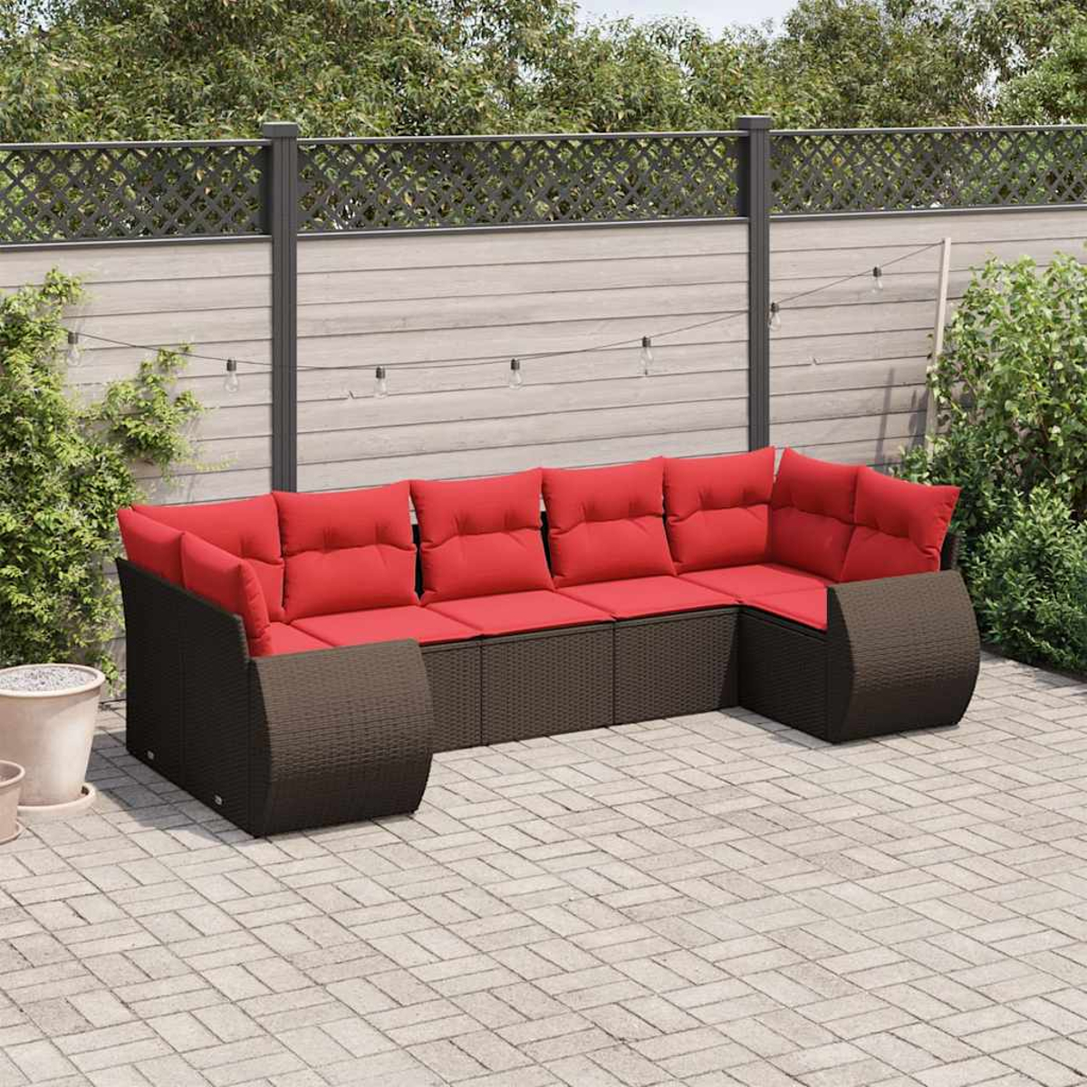 Ein braunes Korbsessel-Sofa mit roten Kissen steht auf einer Backstein-Terrasse. Ein Zaun und Grünzeug sind im Hintergrund.