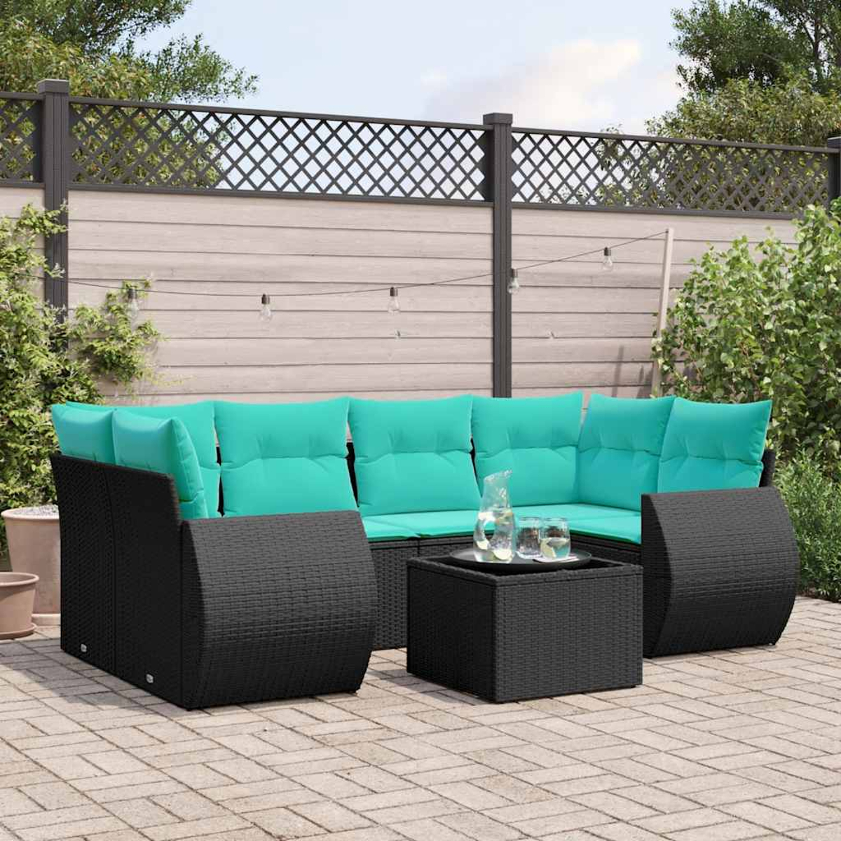 Outdoor-Korbsofa mit türkisfarbenen Kissen, kleinem Tisch und einem Glas Wasser auf einer Ziegelterrasse, im Hintergrund ein Zaun.