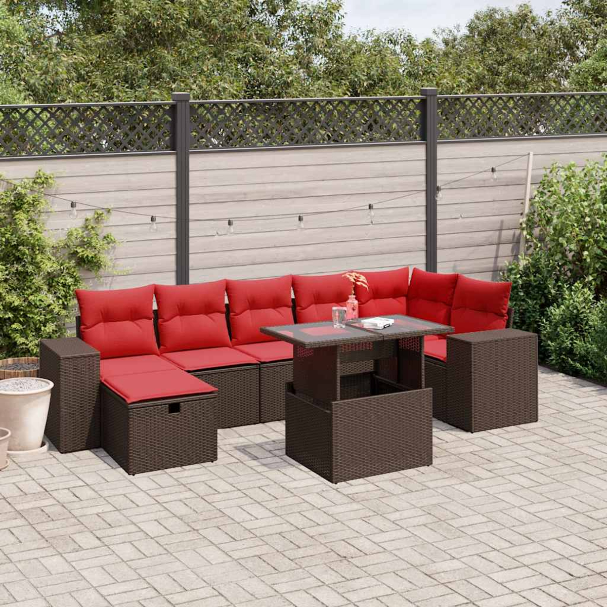 Garten-Set mit roten Kissen, braunen Korbmöbeln und einem Tisch mit Glasplatte. Der Hintergrund zeigt einen Holzzaun und Grün.