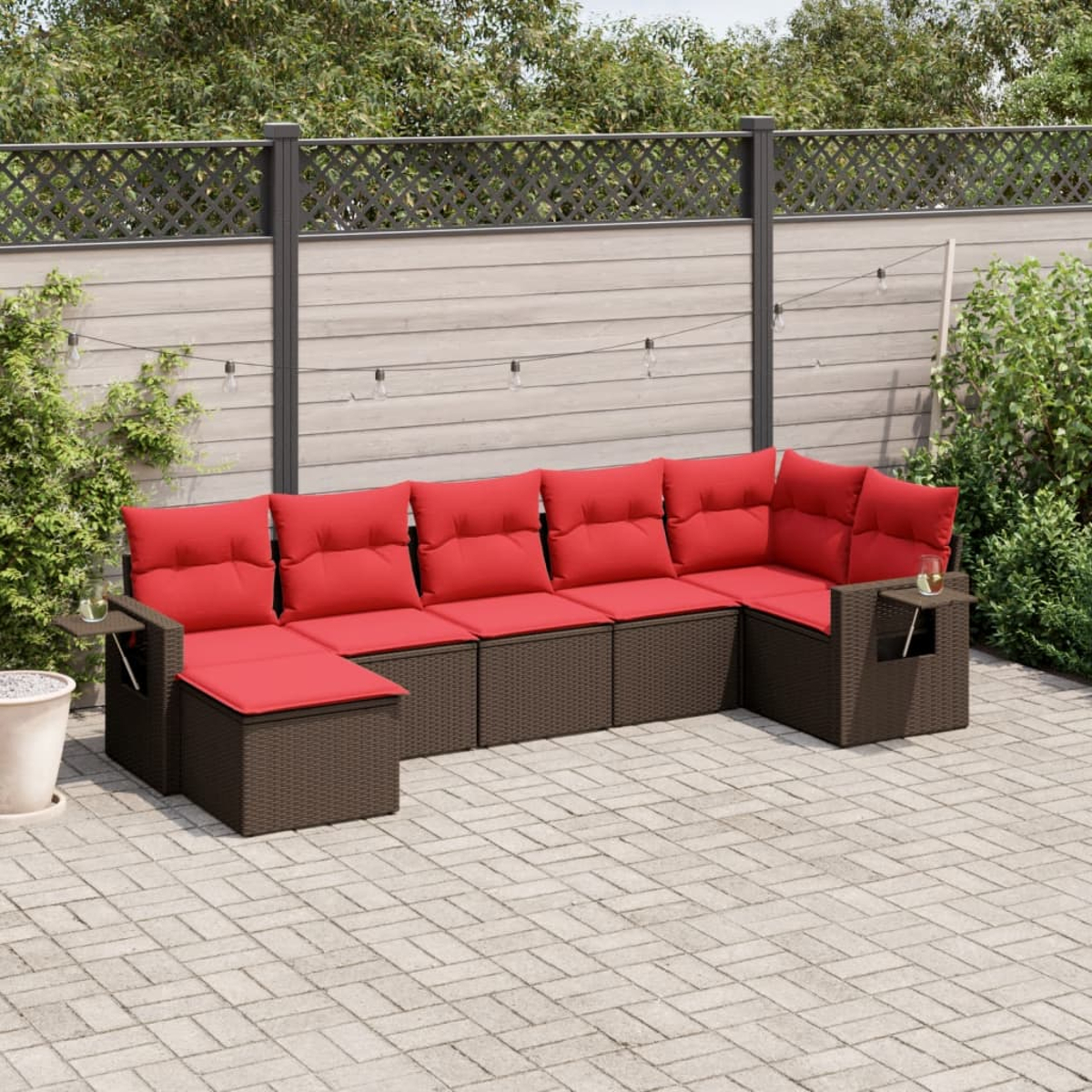 Outdoor-Ecksofa mit roten Kissen, braunem Korbgewebe und Beistelltischen. Auf einer Steinterrasse mit Zaun und Grün im Hintergrund.