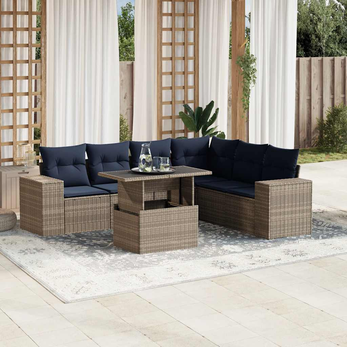 Outdoor-Korbmöbel mit marineblauen Kissen. Ein Tisch mit Glasplatte trägt eine Kanne und Gläser. Hintergrund mit Vorhängen und Holzzaun.
