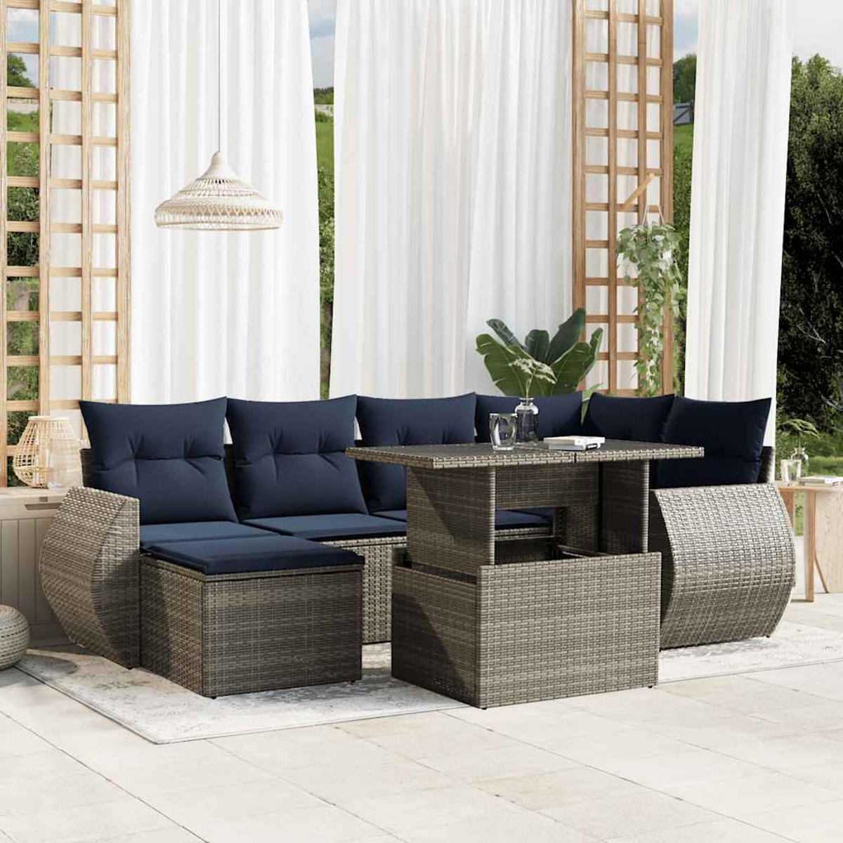 Terrassen-Set mit Tisch, Sofa und Ottomane. Marineblaue Kissen kontrastieren mit grauem Korbgeflecht. Weiße Vorhänge und Pflanzen im Hintergrund.