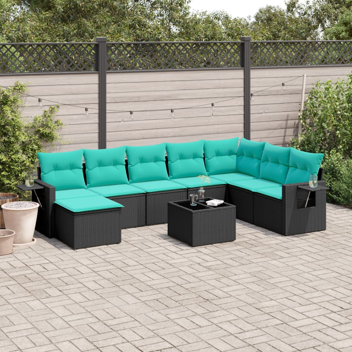 Ein schwarzes Korbsessel-Sofa mit türkisfarbenen Kissen steht auf einer Ziegelterrasse. Davor steht ein kleiner schwarzer Tisch. Hinten ist ein Holzzaun.