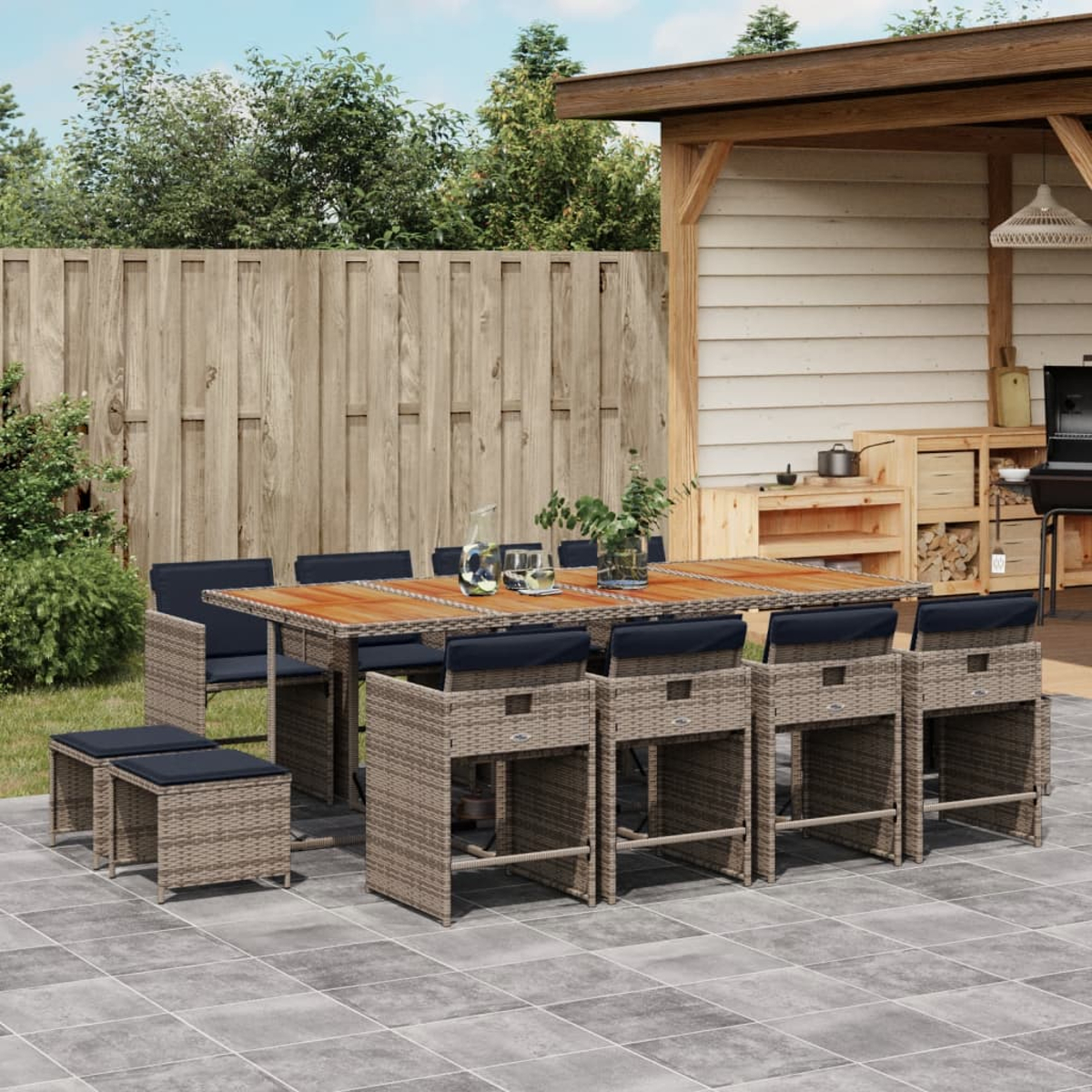 Essgruppe im Freien mit Tisch, Stühlen und Hockern. Holzzaun und Pergola im Hintergrund. Getränke auf dem Tisch, sonniger Tag.