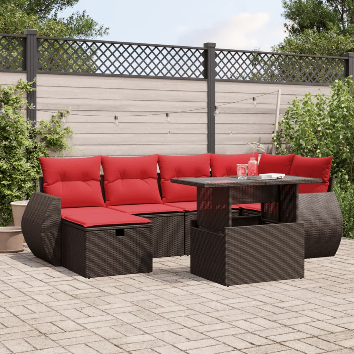 Ein Terrassenset mit einem braunen Korbmöbel-Sofa und -Tisch mit roten Kissen. Im Hintergrund ein Zaun und Pflanzen.