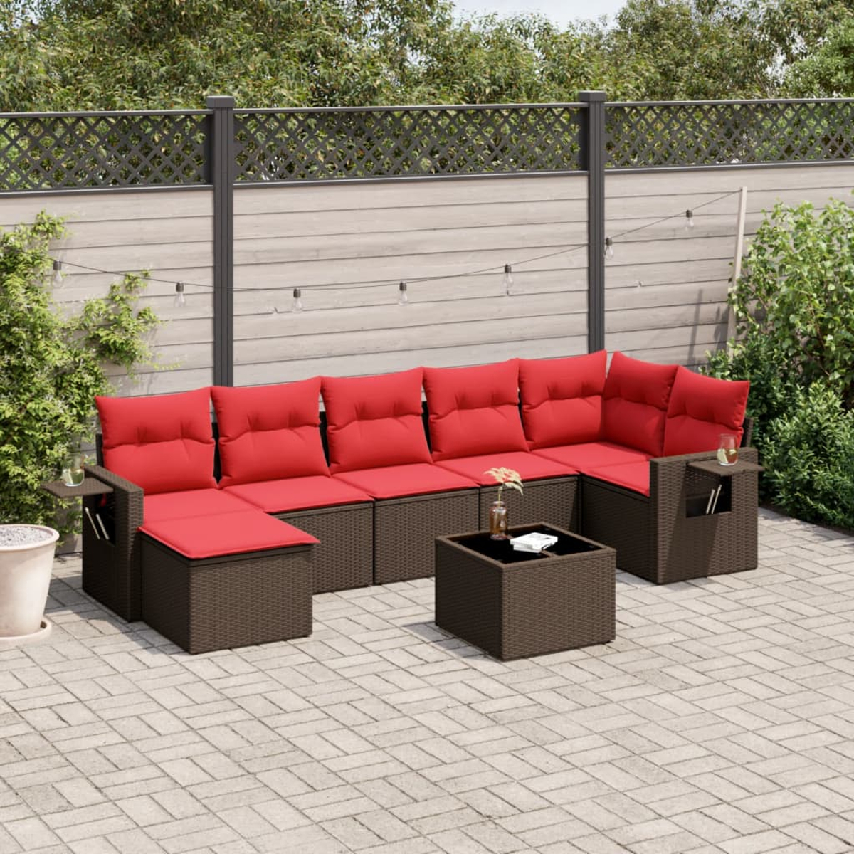 Outdoor-Sofa mit roten Kissen und braunem Korbgeflecht. Davor steht ein kleiner Tisch. Im Hintergrund befinden sich ein Zaun und Pflanzen.