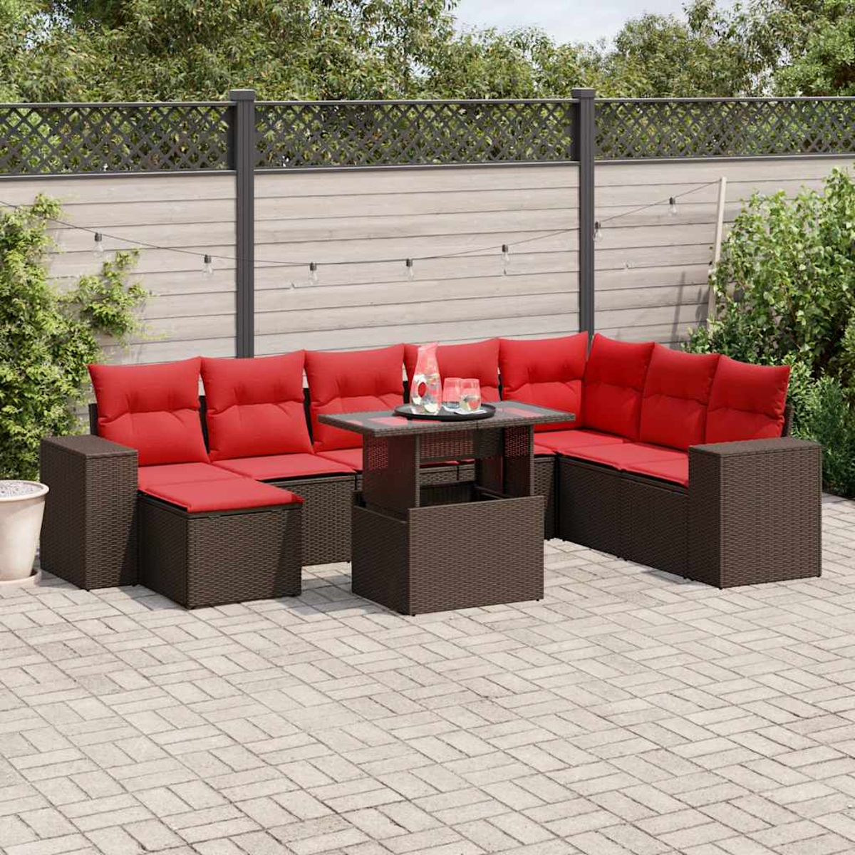 Juego de asientos al aire libre con cojines rojos y una mesa. Muebles de mimbre marrón en un patio de piedra, rodeado de una valla y vegetación.