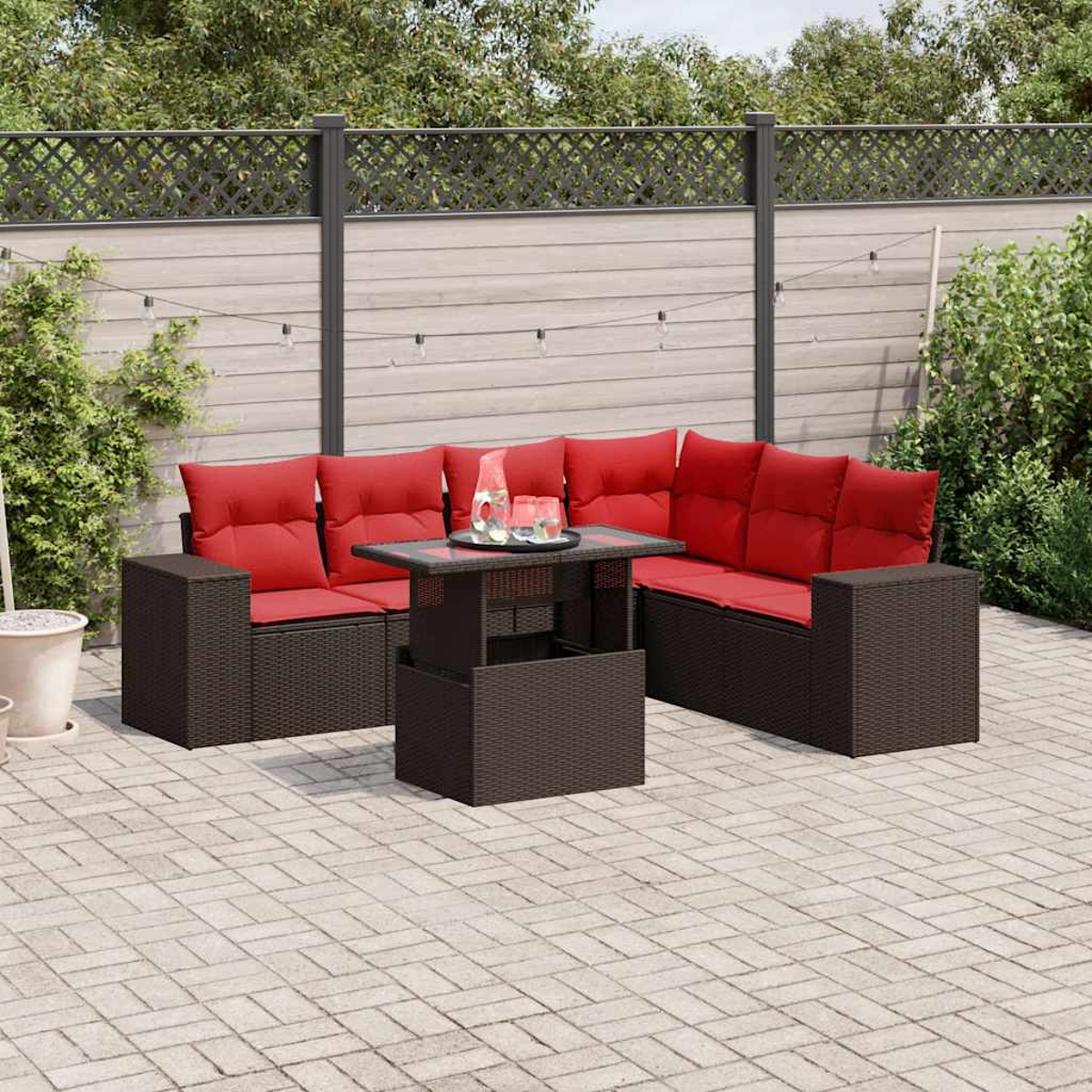 Garten-Terrassenset mit roten Kissen, Glastisch und dunkelbraunem Korbgeflecht. Das Set steht auf einer Ziegelterrasse. Im Hintergrund ein Holzzaun.