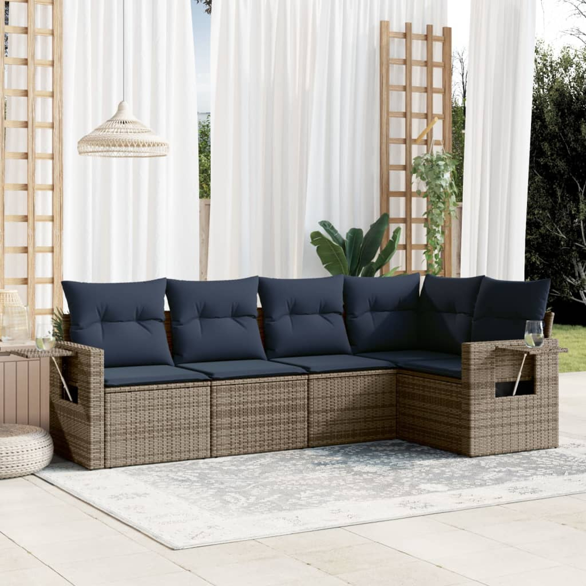 Ein Ecksofa aus Korbgeflecht mit marineblauen Kissen steht auf einem Teppich auf der Terrasse. Weiße Vorhänge und ein Spalier im Hintergrund.