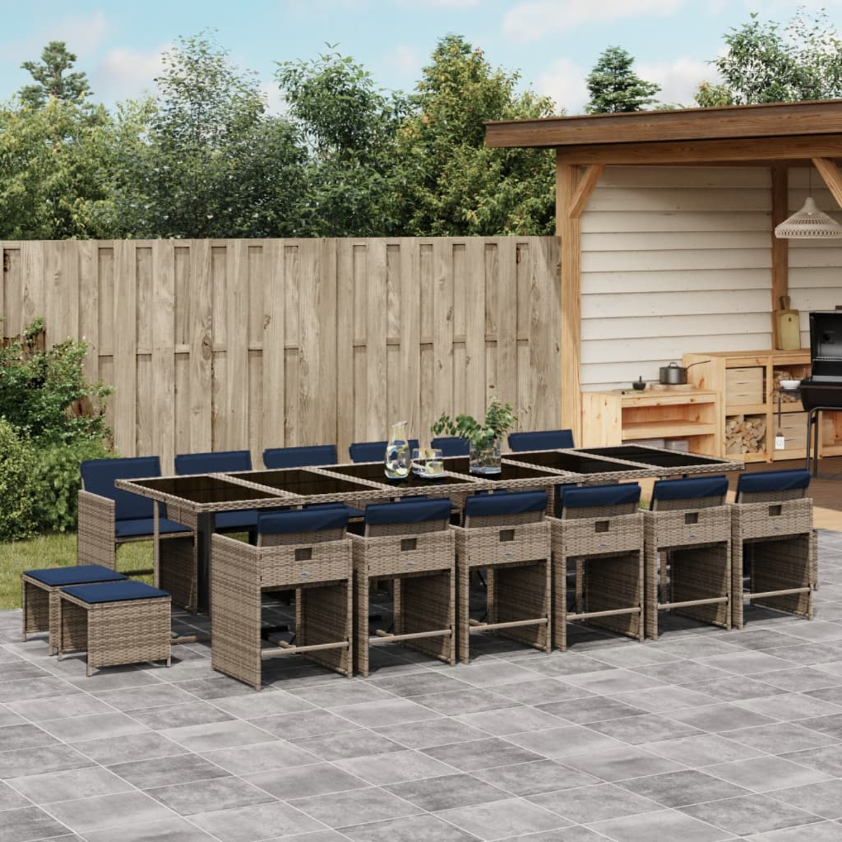 Essgruppe im Freien. Rattantisch mit Stühlen und blauen Kissen. Terrasse mit Holzzaun und Bäumen im Hintergrund.