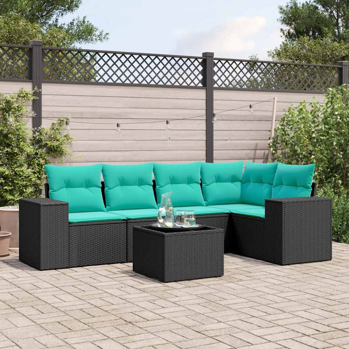 Schwarze Outdoor-Korbmöbel mit türkisfarbenen Kissen. Ein Tisch mit Krug und Gläsern steht auf den Terrassensteinen. Zaun und Pflanzen im Hintergrund.