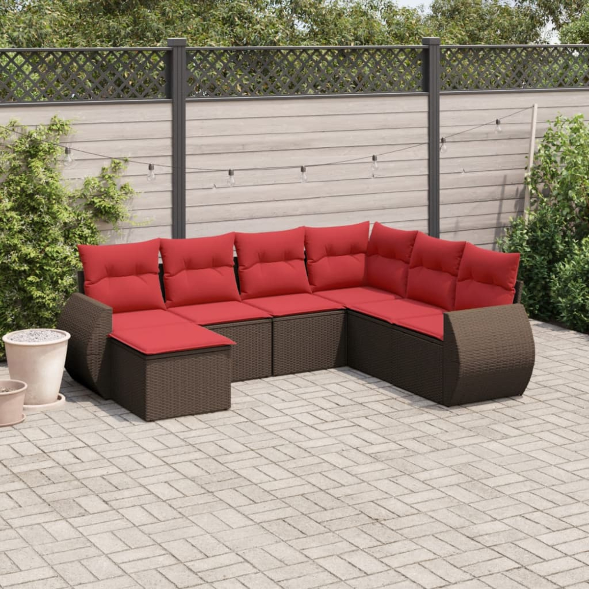 Ein Ecksofa im Freien mit roten Kissen steht auf einer Terrasse. Ein Zaun und Grünzeug sind im Hintergrund. Lichterketten sind sichtbar.