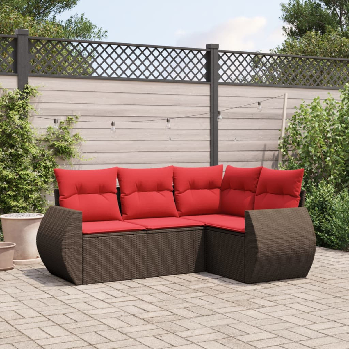 Ein braunes Ecksofa aus Korbgeflecht mit roten Kissen steht auf einer Ziegelterrasse. Hintergrund: Zaun, Pflanzen und Lichterkette.