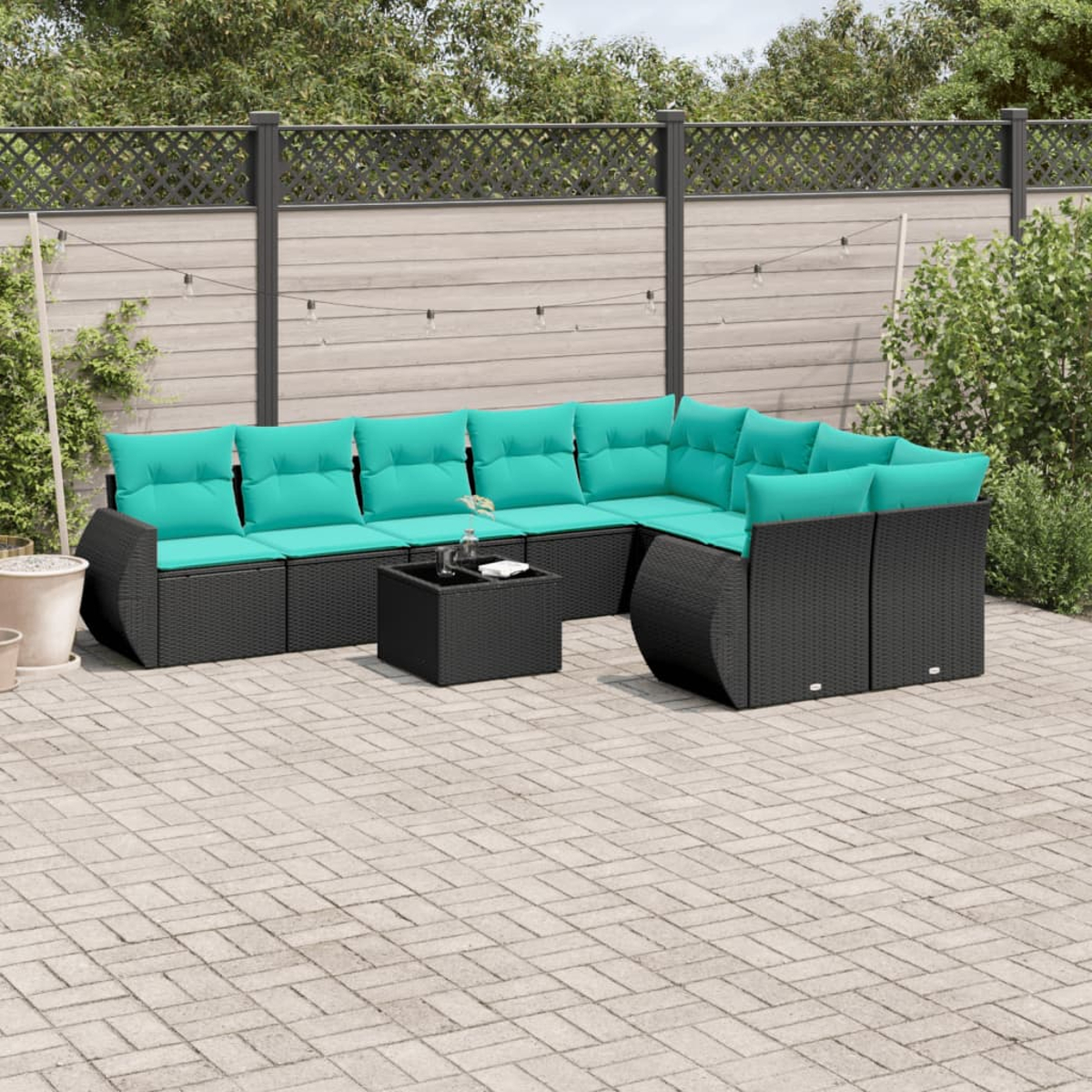 Ein schwarzes Korbsessel-Ecksofa mit tealfarbenen Kissen und einem kleinen Tisch steht auf einer Ziegelterrasse, vor einem Zaun und Grün.