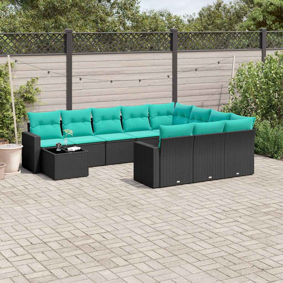 Ein schwarzes Korbsessel-Sofa mit tealfarbenen Kissen steht auf einer Ziegelterrasse. Ein kleiner Tisch mit Getränken. Ein Zaun und Pflanzen im Hintergrund.