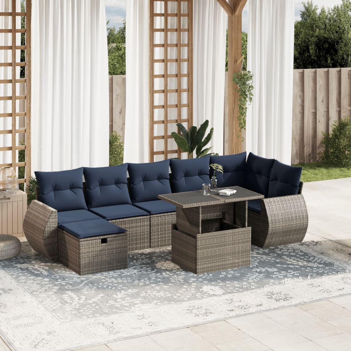 Garten-Sitzgruppe mit blauen Kissen und einem Tisch mit Hubfunktion auf einem gemusterten Teppich. Durchsichtige Vorhänge und Grün im Hintergrund.