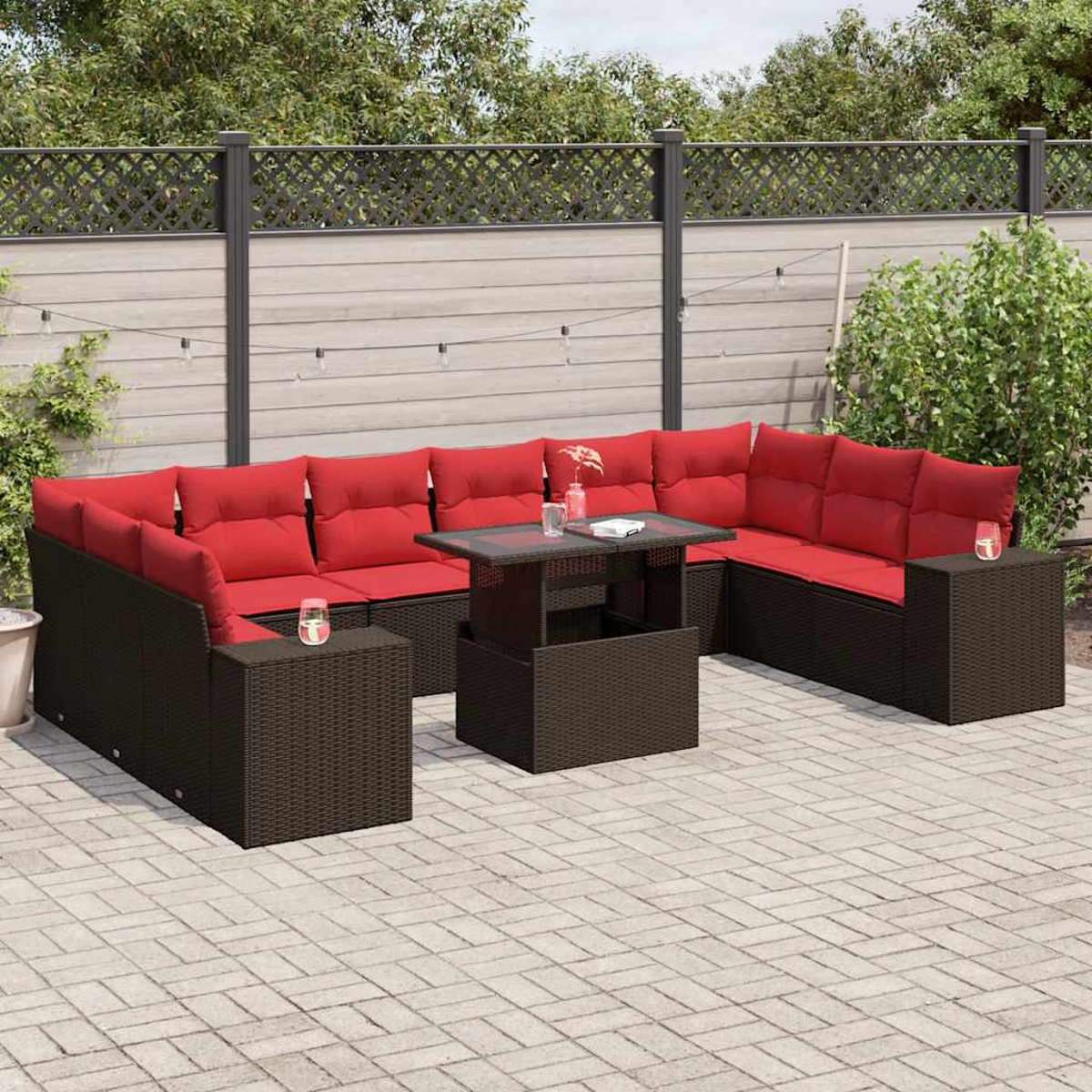 Terrassenset mit roten Kissen, braunen Korbmöbeln, Glastisch und Beistelltischen. Hintergrund: Zaun, Grün und Backstein-Patio.