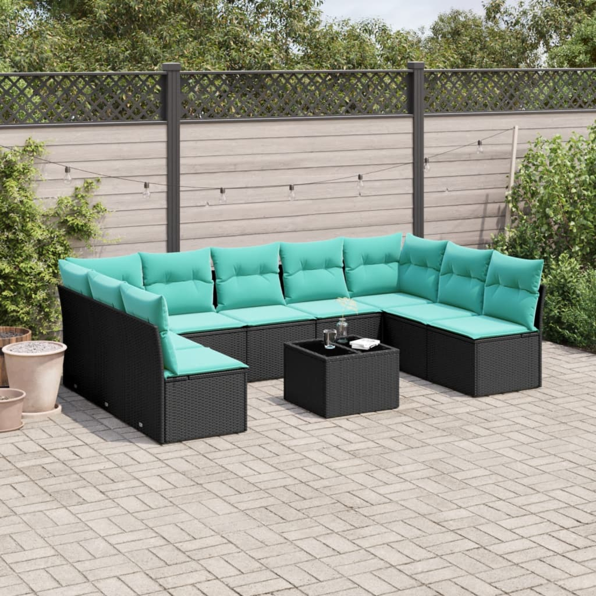 Ein schwarzes Korbsessel-Sofa mit türkisfarbenen Kissen steht auf einer Backstein-Terrasse. Davor steht ein quadratischer schwarzer Couchtisch. Im Hintergrund ist ein Zaun.