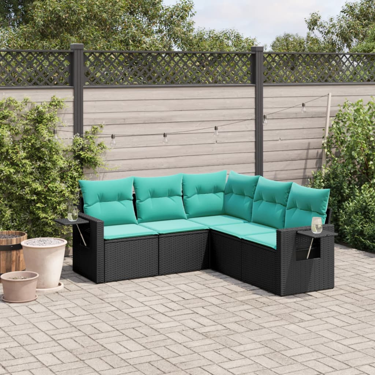 Schwarzes Korbsessel-Sofa mit türkisfarbenen Kissen auf einer Backstein-Terrasse. Im Hintergrund ein Holzzaun, Pflanzen und dekorative Lichter.