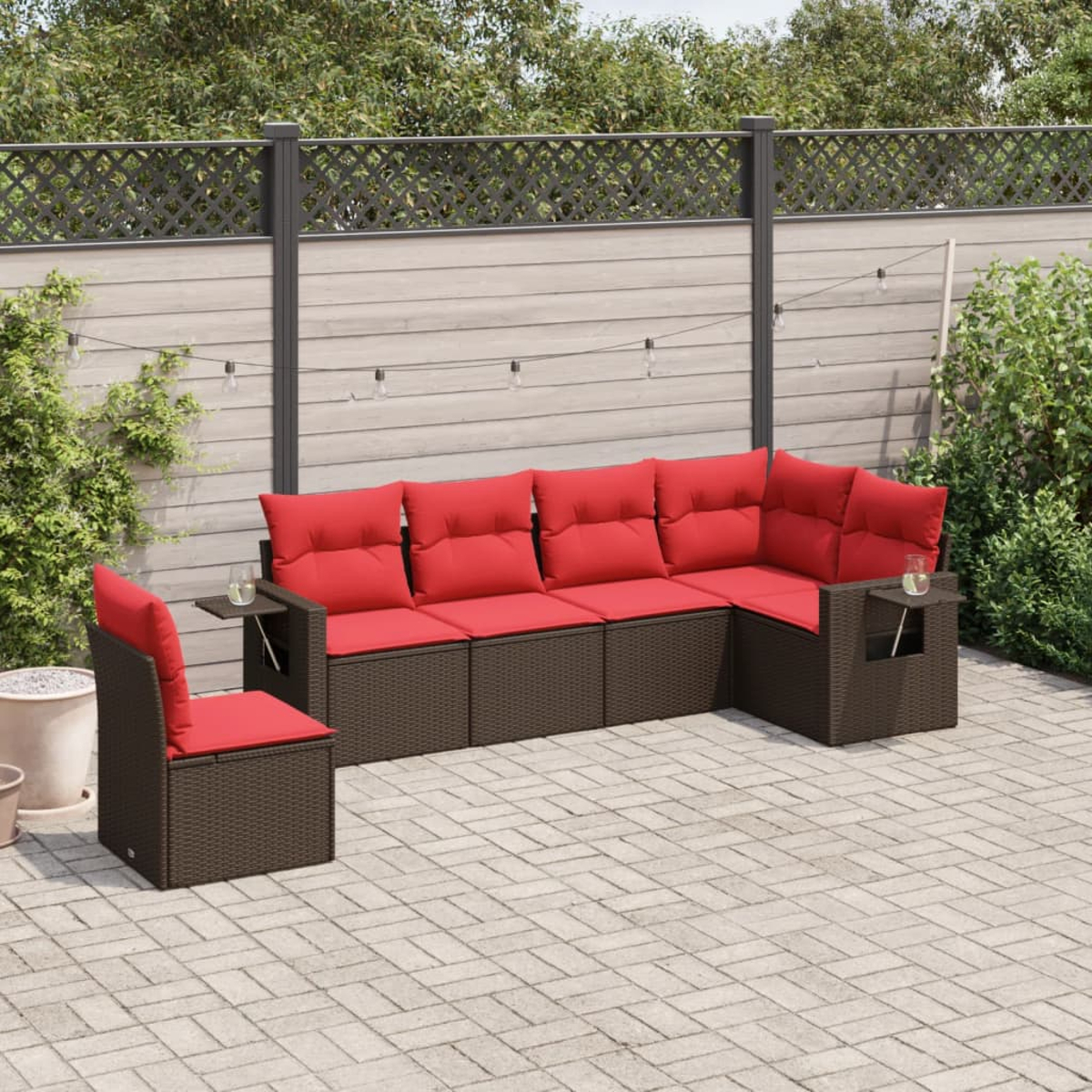 Ein Ecksofa für draußen mit roten Kissen und braunem Korbgeweberahmen, ein Stuhl und eine Backstein-Terrasse. Der Hintergrund zeigt einen Zaun und Grün.