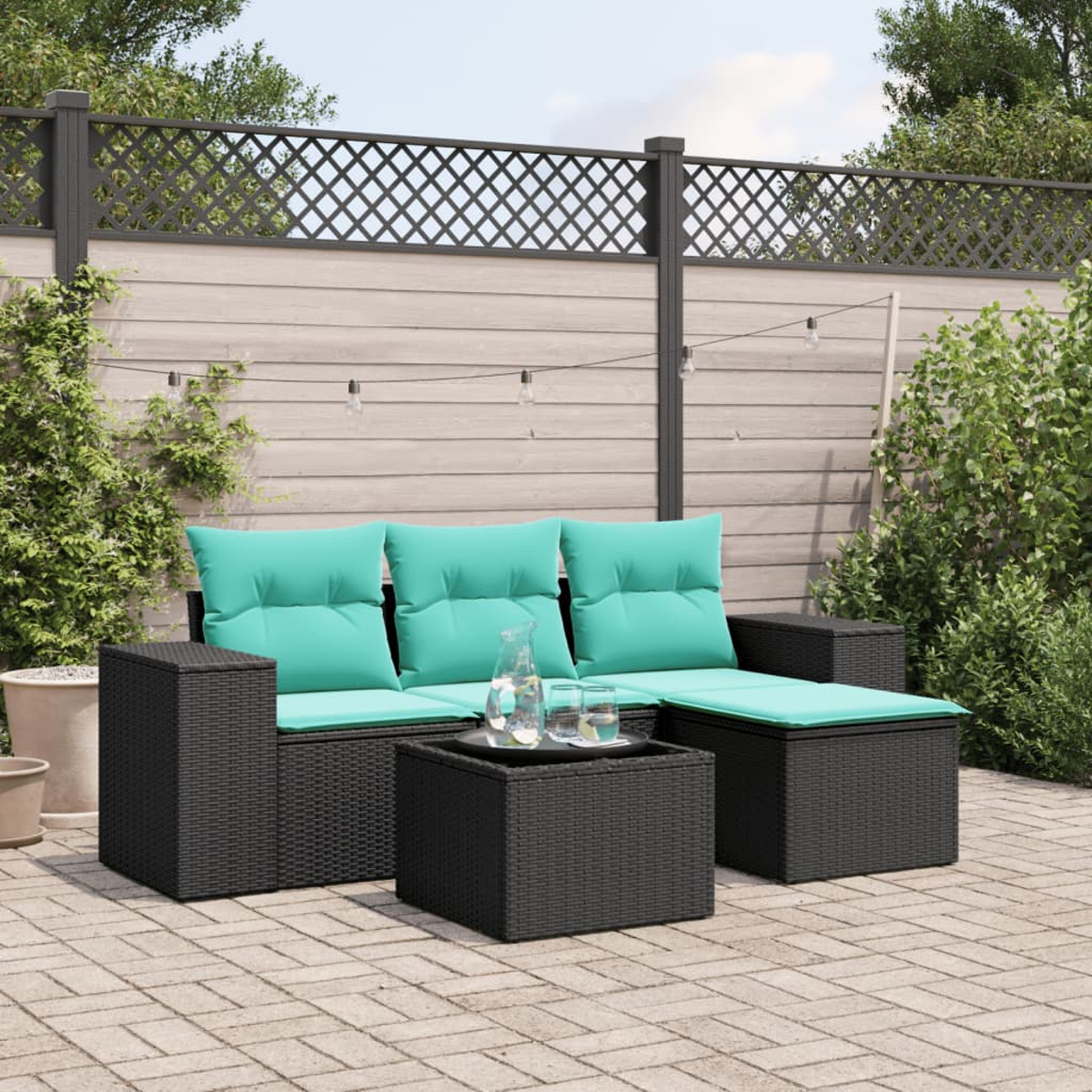 Garten-Lounge-Set mit schwarzem Korbmöbel und teal-farbenen Kissen. Besteht aus Sofa, Ottomane und Couchtisch. Hintergrund mit Zaun und Grün.