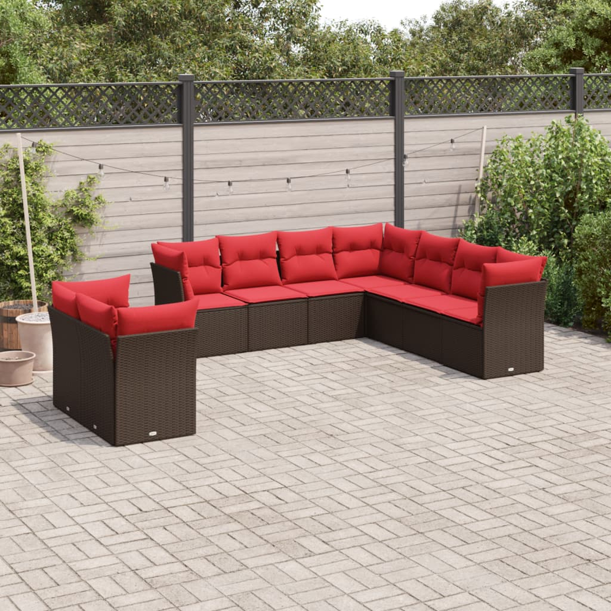 Outdoor-Ecksofa mit roten Kissen auf einer gepflasterten Terrasse. Braune Korbmöbel. Hintergrund: Zaun, Pflanzen und Lichterketten.