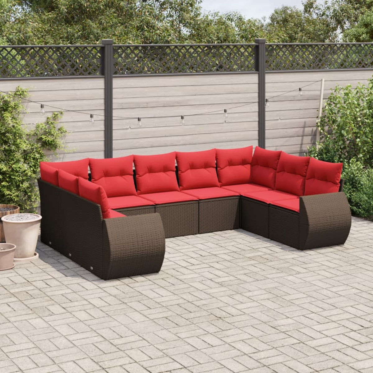 Ein Ecksofa für den Außenbereich mit roten Kissen. Es ist braunes Geflecht auf einer Ziegelterrasse. Ein Zaun, Pflanzen und Lichterketten sind im Hintergrund.