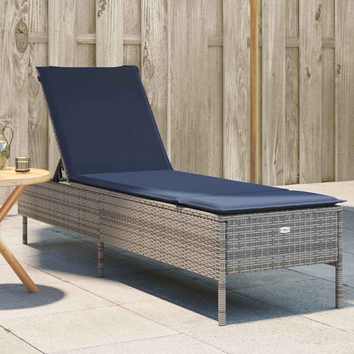 Eine graue Korbliege mit marineblauem Kissen steht auf einer Terrasse. Links steht ein kleiner Tisch mit einem Krug. Dahinter ein Holzzaun.