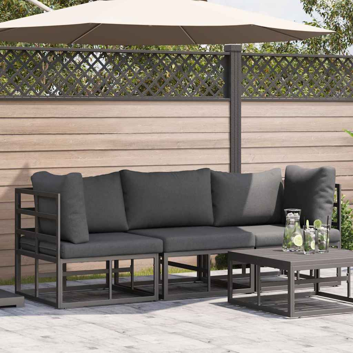 Set da patio esterno con cuscini grigi, un tavolino e bevande. Un grande ombrellone fornisce ombra. Recinzione in legno sullo sfondo.