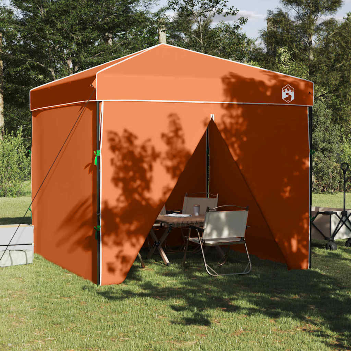 Ein orangefarbenes Zelt steht auf Gras. Im Inneren sind ein Tisch und Stühle aufgestellt. Bäume und Sonnenlicht werfen Schatten auf Zelt und Boden.