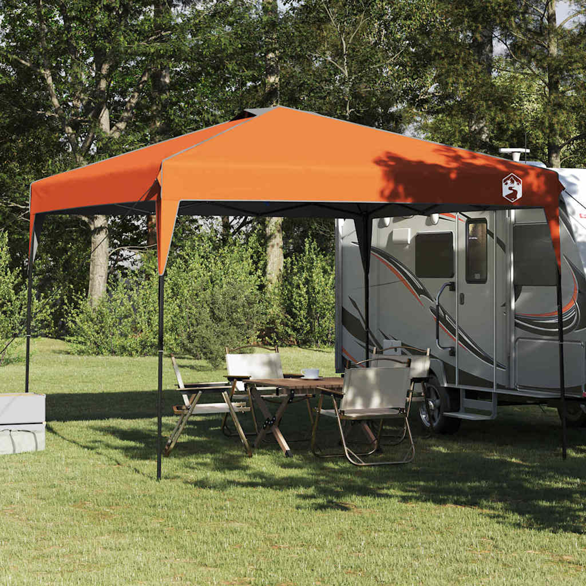 Una tenda arancione ombreggia un campeggio. Un rimorchio è parcheggiato nelle vicinanze, con sedie e un tavolo sull'erba. Gli alberi sono sullo sfondo.