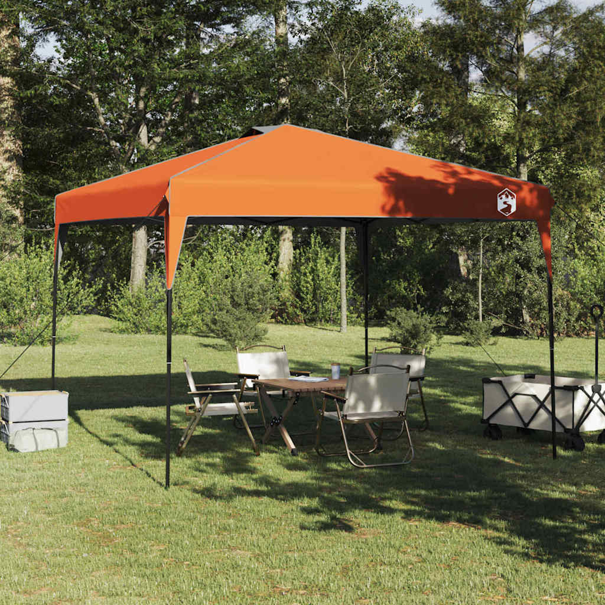 Una tenda arancione sopra un tavolo e delle sedie su un prato. Due carrelli e alberi sullo sfondo.