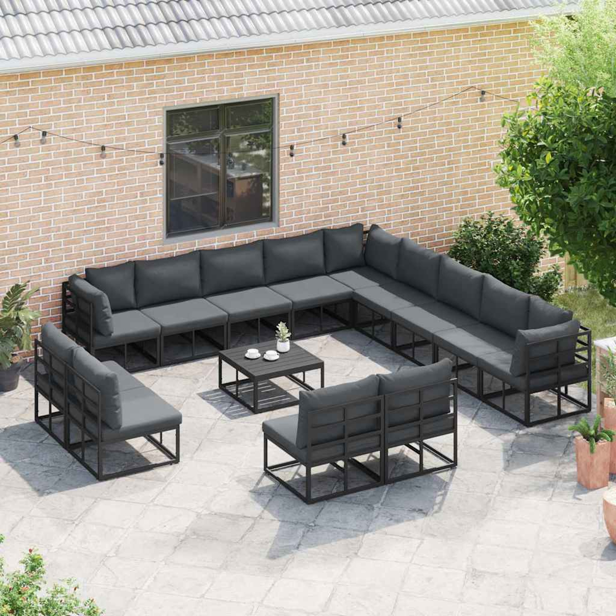 Outdoor-Sitzbereich mit grauen Kissen, einem schwarzen Tisch und Topfpflanzen. Ziegelmauer im Hintergrund und Lichterketten.