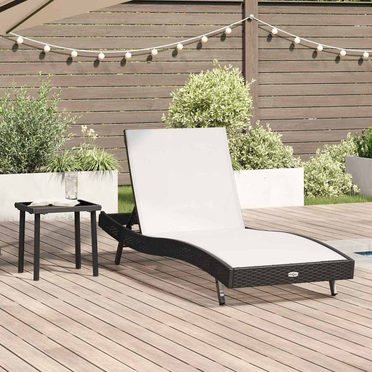 Szene im Freien mit einer schwarzen Liege mit weißem Kissen auf einem Holzdeck, kleinem Tisch, Pflanzen und Lichterketten.