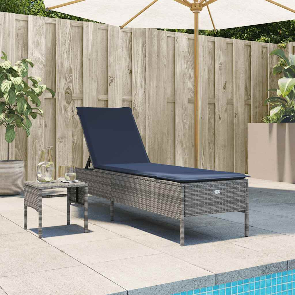 Una tumbona de mimbre gris con un cojín azul está cerca de una mesita, una jarra de agua y una sombrilla junto a una piscina.